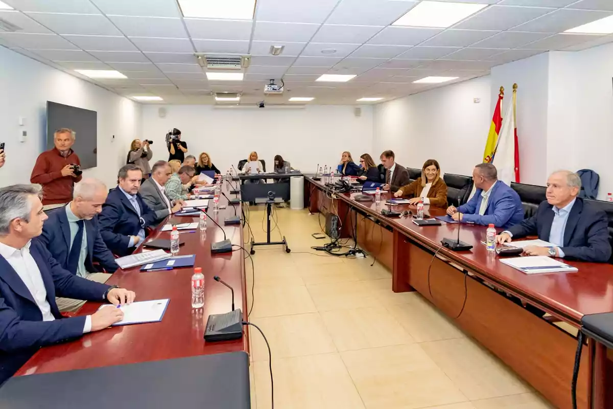 Varias personas sentadas alrededor de una mesa de reuniones en una sala de conferencias, algunas con carpetas y botellas de agua, mientras otras personas toman fotografías al fondo Varias personas sentadas alrededor de una mesa de reuniones en una sala de conferencias, algunas con carpetas y botellas de agua, mientras otras personas toman fotografías al fondo