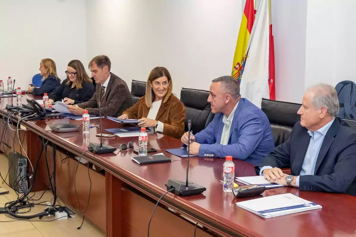 Varias personas sentadas en una mesa de reuniones con documentos y botellas de agua, conversando y trabajando en un ambiente formal con banderas al fondo.