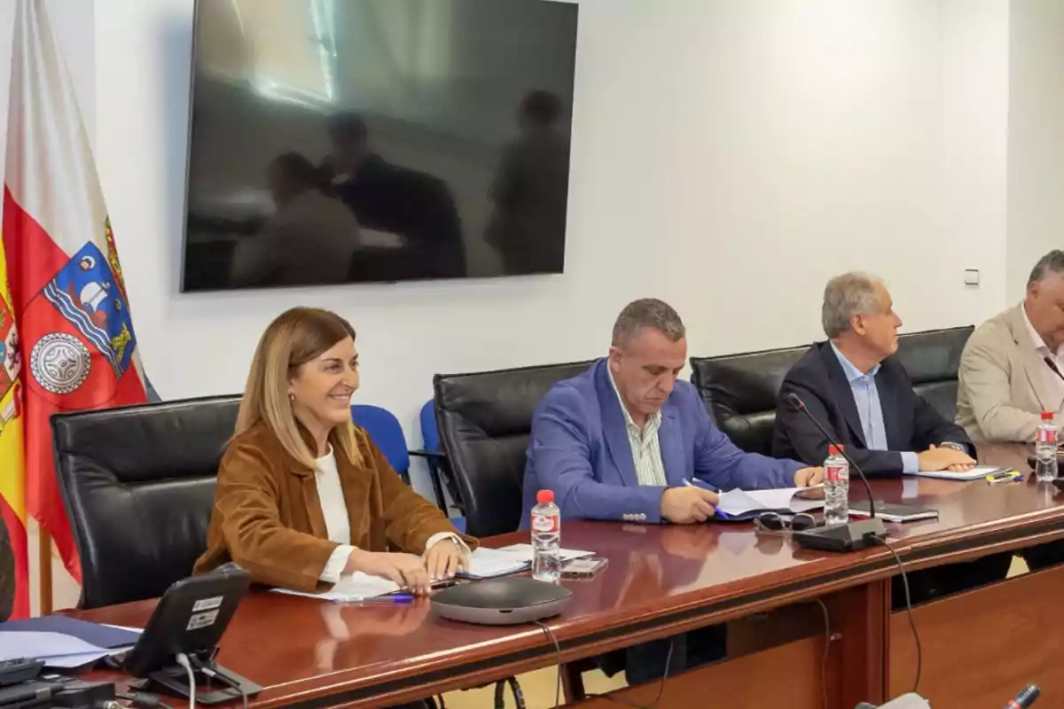Cuatro personas sentadas en una mesa de reuniones con documentos y botellas de agua, al fondo hay una pantalla grande y banderas oficiales. Cuatro personas sentadas en una mesa de reuniones con documentos y botellas de agua, al fondo hay una pantalla grande y banderas oficiales.
