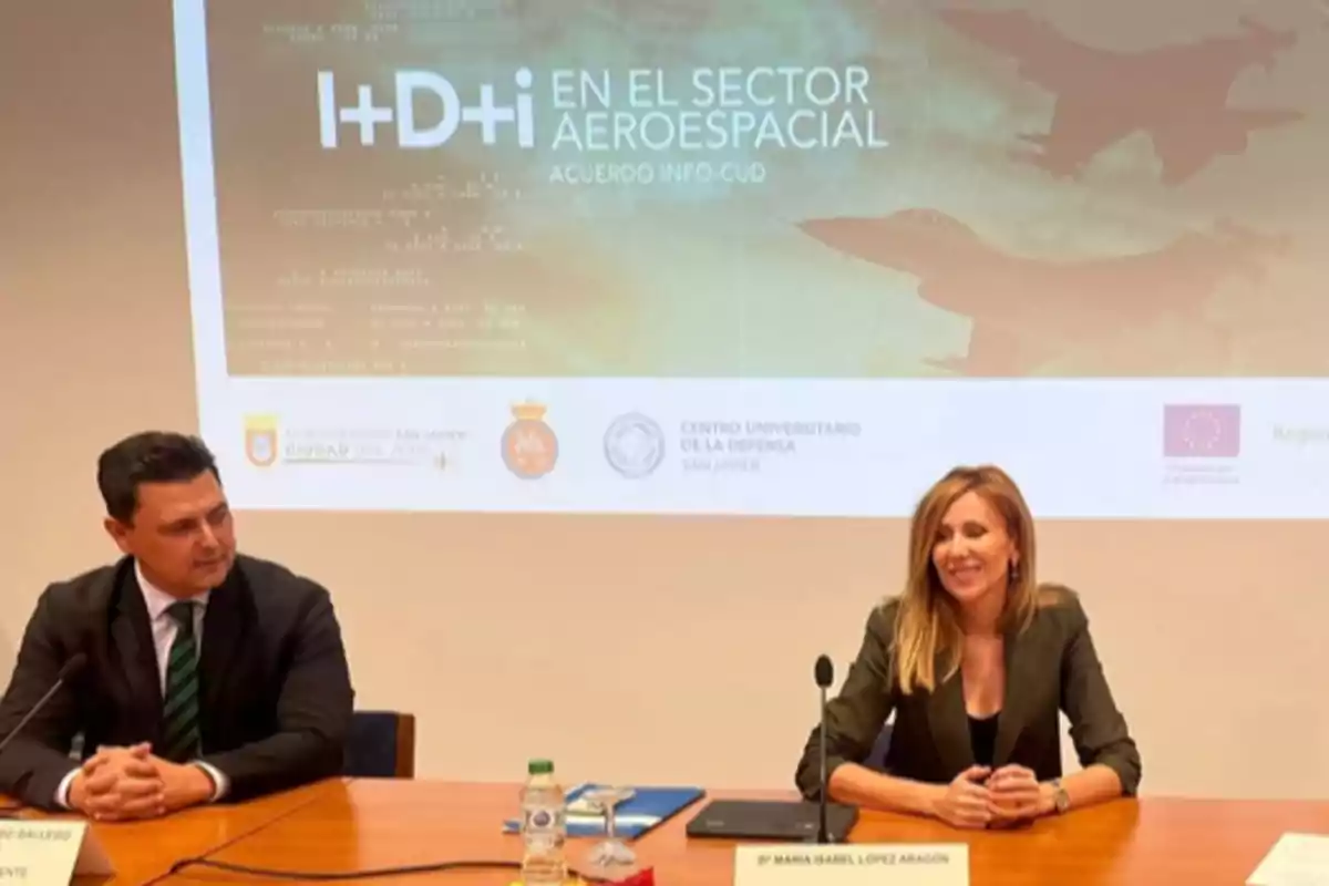 Dos personas sentadas en una mesa durante una conferencia sobre innovación y desarrollo en el sector aeroespacial con una pantalla de presentación al fondo Dos personas sentadas en una mesa durante una conferencia sobre innovación y desarrollo en el sector aeroespacial con una pantalla de presentación al fondo