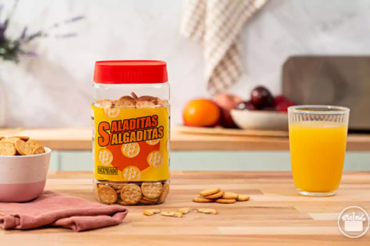 Un frasco de galletas saladas junto a un vaso de jugo de naranja y un tazón pequeño con galletas sobre una mesa de madera en una cocina