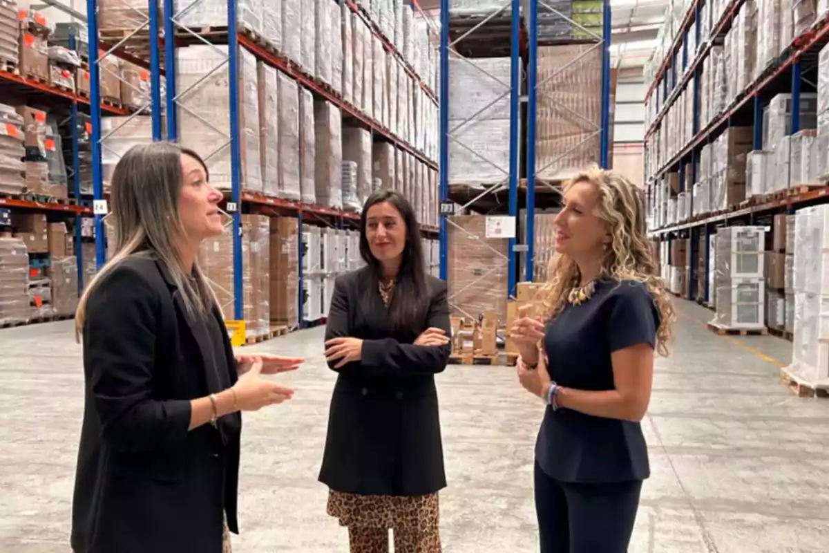 Tres mujeres conversan en el interior de un almacén industrial rodeadas de estanterías con cajas y mercancía empaquetada