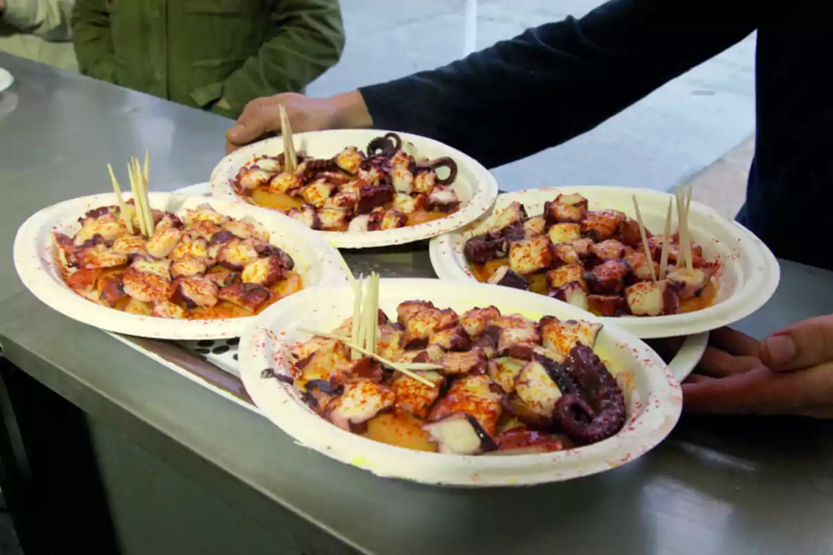 Cuatro platos de pulpo a la gallega servidos en platos desechables con palillos sobre una mesa de metal