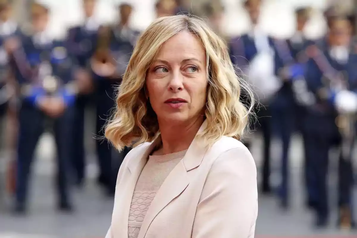 Mujer de cabello rubio y ondulado con expresión seria, vestida con saco claro, de pie frente a un grupo de personas uniformadas desenfocadas al fondo Mujer de cabello rubio y ondulado con expresión seria, vestida con saco claro, de pie frente a un grupo de personas uniformadas desenfocadas al fondo