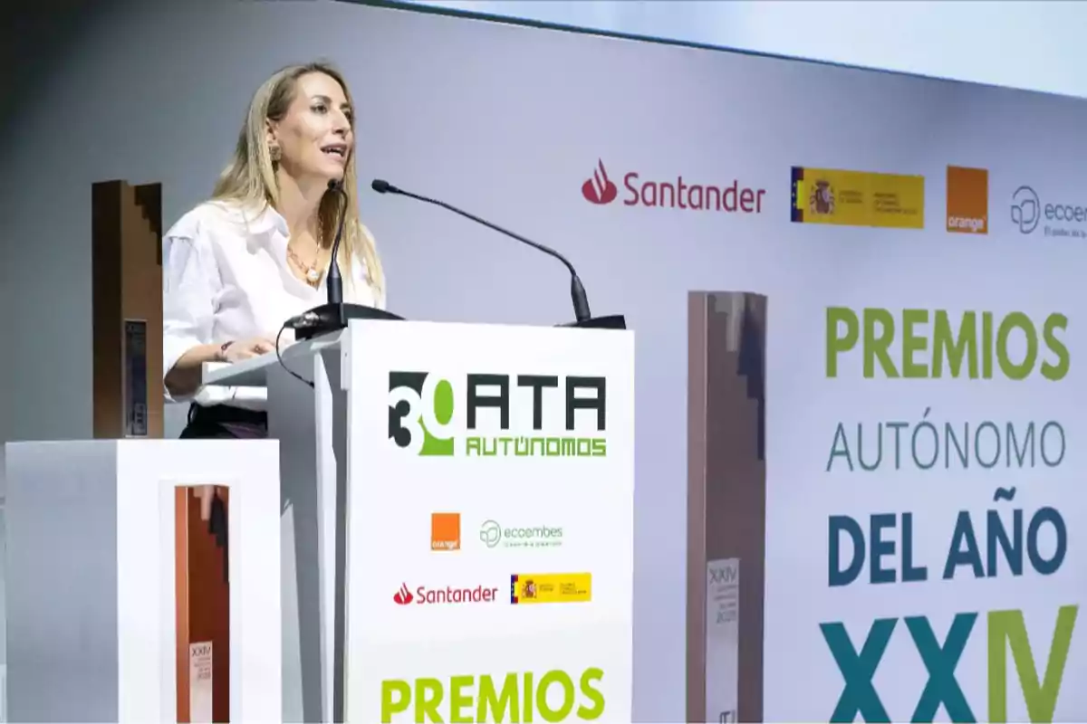 Mujer rubia hablando en un podio durante la ceremonia de los Premios Autónomo del Año organizada por ATA Autónomos con logotipos de patrocinadores y trofeos visibles en el escenario