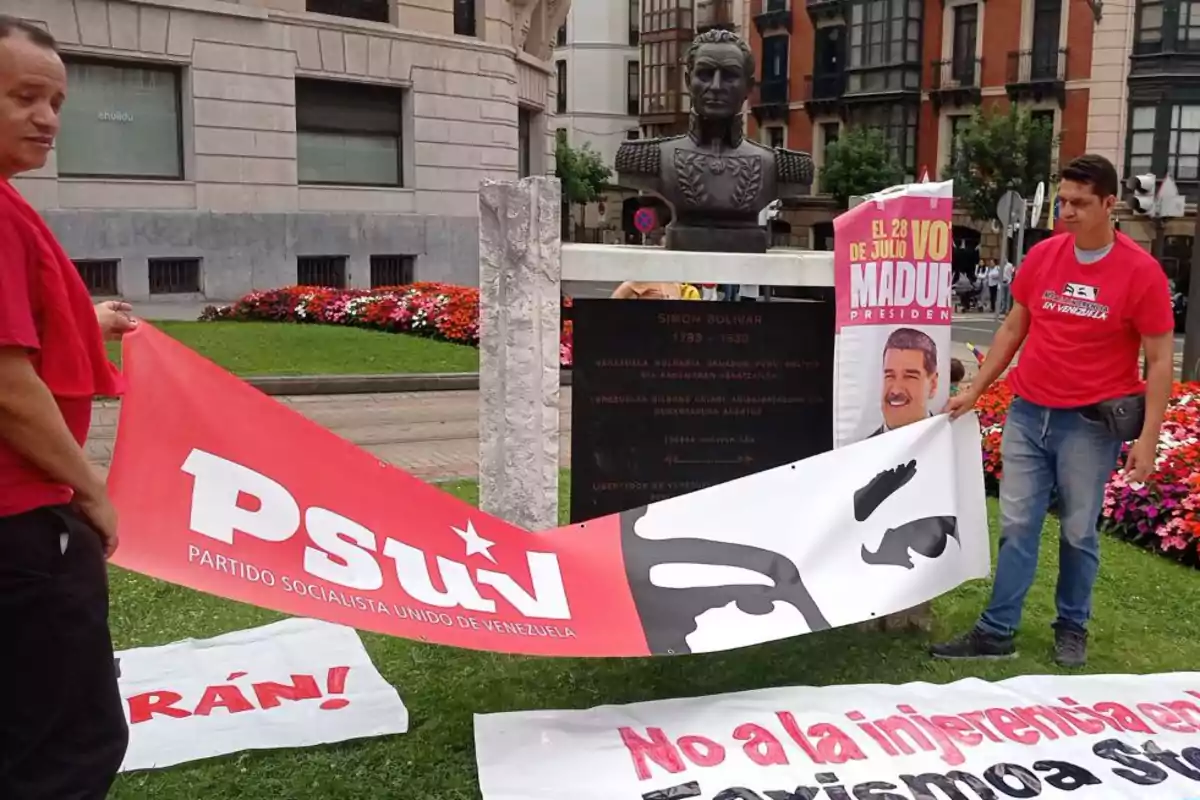 Dos personas sostienen una pancarta del Partido Socialista Unido de Venezuela frente a un busto de Simón Bolívar, junto a carteles de apoyo a Nicolás Maduro y mensajes políticos en un espacio público con flores y edificios al fondo Dos personas sostienen una pancarta del Partido Socialista Unido de Venezuela frente a un busto de Simón Bolívar, junto a carteles de apoyo a Nicolás Maduro y mensajes políticos en un espacio público con flores y edificios al fondo