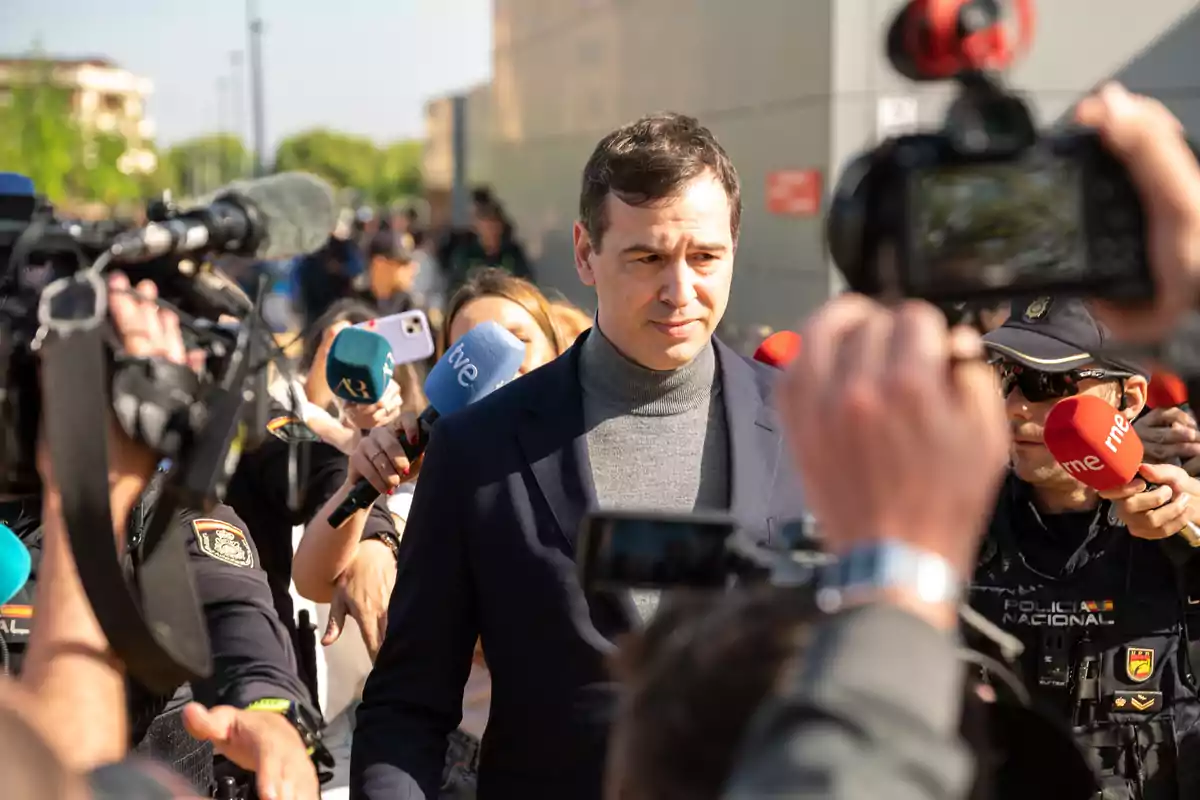 Un hombre con suéter gris y saco oscuro es rodeado por periodistas y cámaras mientras responde preguntas en la calle, acompañado de policías