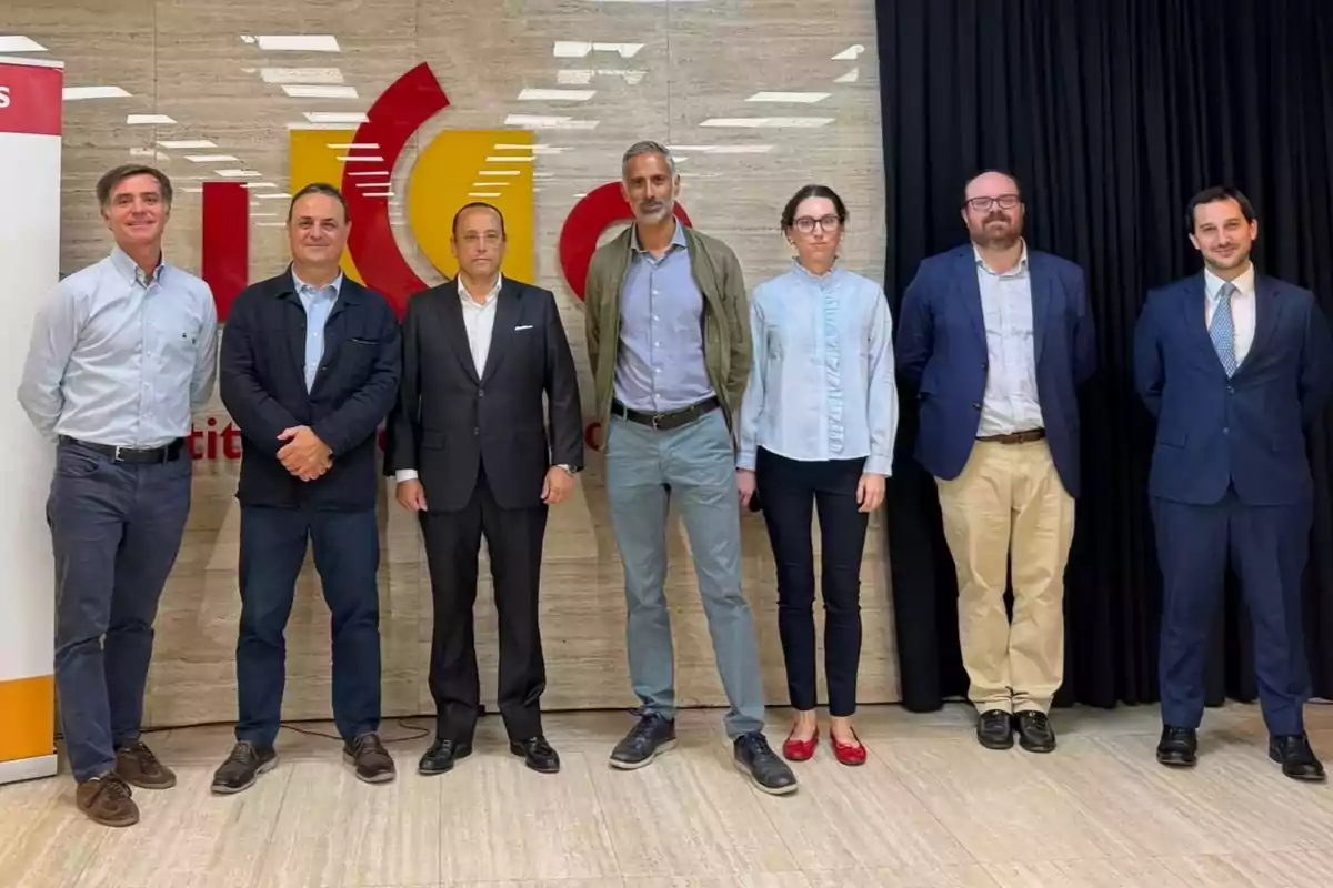 Siete personas posan de pie en fila frente a una pared con un logotipo color rojo y amarillo, todas vestidas de manera formal y casual, en un entorno interior iluminado.