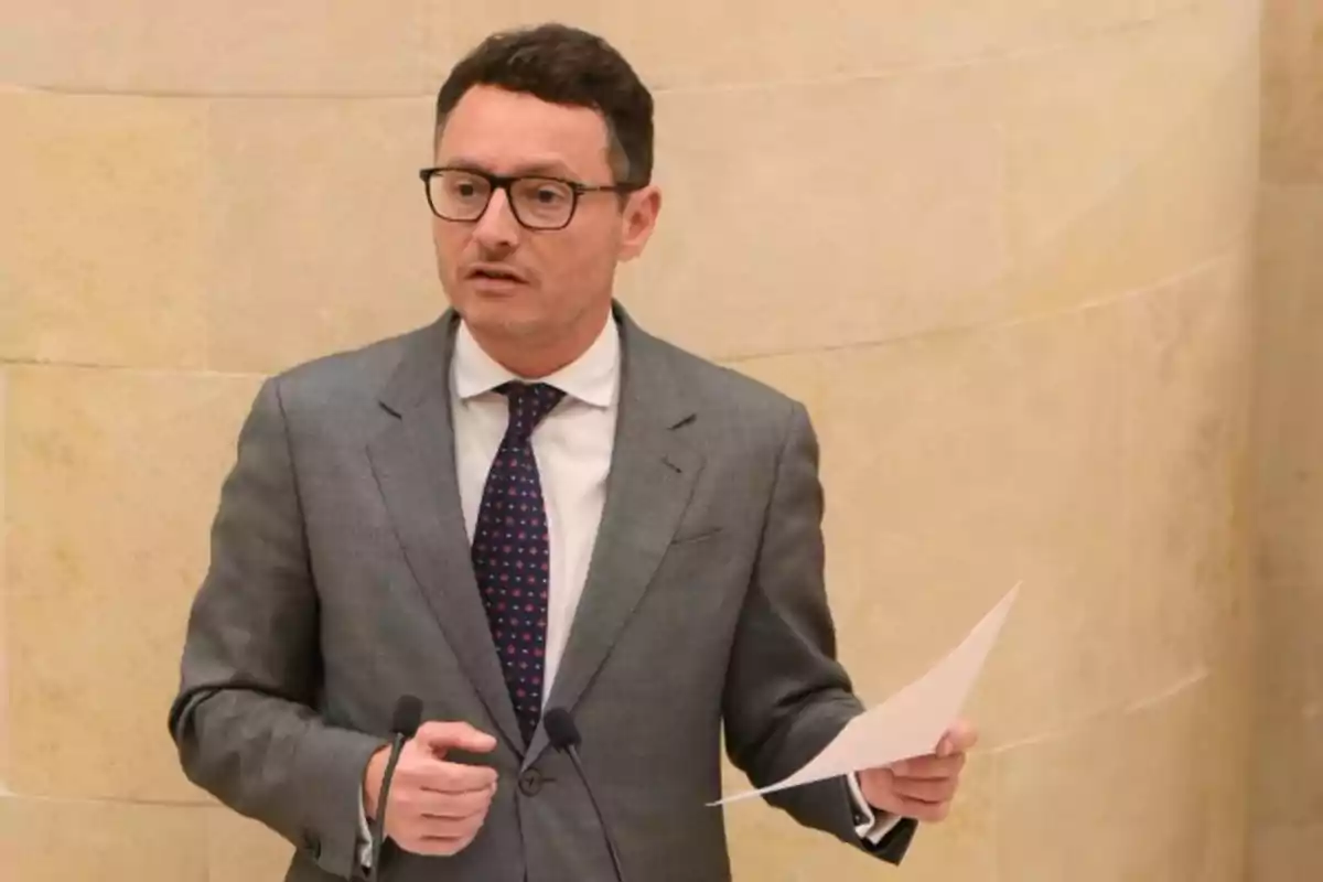 Hombre con gafas y traje gris hablando frente a dos micrófonos mientras sostiene una hoja de papel en la mano derecha Hombre con gafas y traje gris hablando frente a dos micrófonos mientras sostiene una hoja de papel en la mano derecha