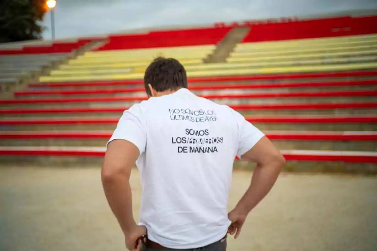 Hombre de espaldas con camiseta blanca que tiene un mensaje motivacional en la espalda, de pie frente a gradas vacías de colores rojo y amarillo. Hombre de espaldas con camiseta blanca que tiene un mensaje motivacional en la espalda, de pie frente a gradas vacías de colores rojo y amarillo.
