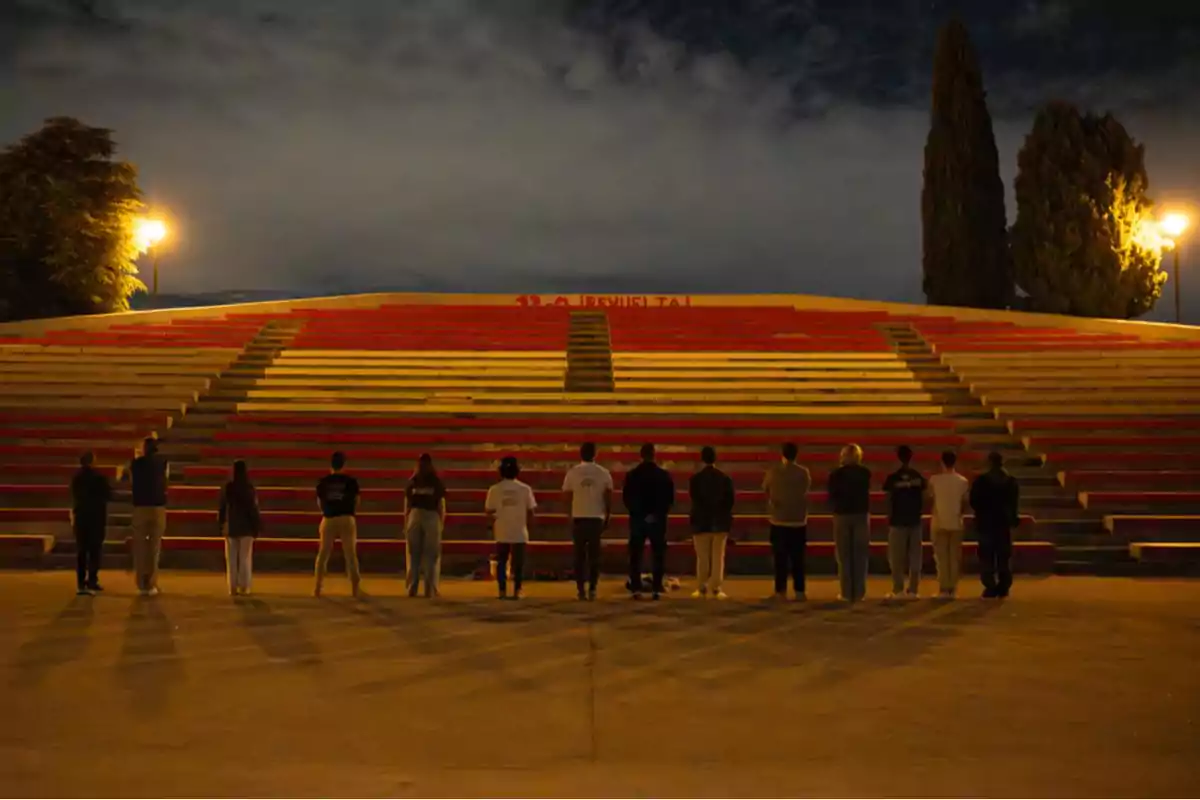 Un grupo de personas de pie en fila frente a unas gradas iluminadas por faroles durante la noche Un grupo de personas de pie en fila frente a unas gradas iluminadas por faroles durante la noche