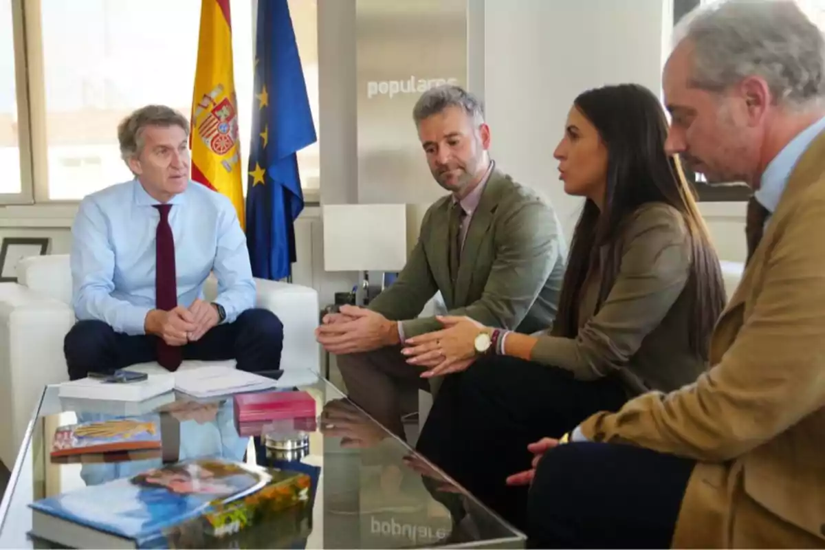 Cuatro personas sentadas en una reunión de oficina con banderas de España y la Unión Europea al fondo y varios documentos sobre la mesa de cristal