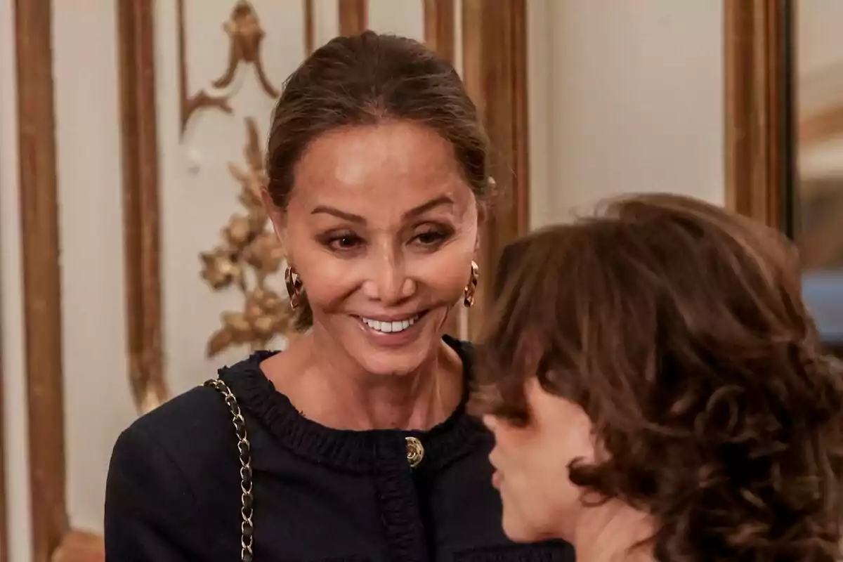 Mujer sonriente con el cabello recogido y vestido oscuro conversando con otra persona en un entorno elegante con detalles dorados en la pared Mujer sonriente con el cabello recogido y vestido oscuro conversando con otra persona en un entorno elegante con detalles dorados en la pared