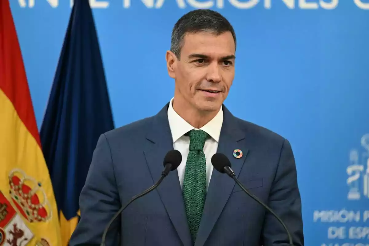Hombre con traje azul y corbata verde hablando frente a micrófonos con una bandera de España al fondo y un fondo azul con texto blanco Hombre con traje azul y corbata verde hablando frente a micrófonos con una bandera de España al fondo y un fondo azul con texto blanco