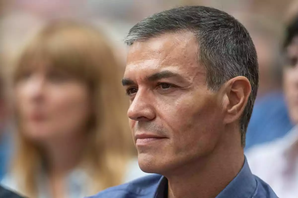 Hombre de cabello corto y canoso con expresión seria en un evento público con personas desenfocadas al fondo Hombre de cabello corto y canoso con expresión seria en un evento público con personas desenfocadas al fondo