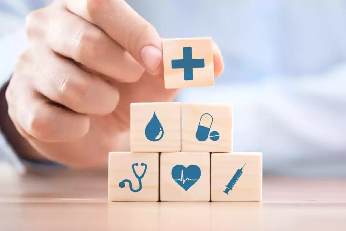 Mano apilando bloques de madera con iconos relacionados con la salud como una cruz, una gota, pastillas, un estetoscopio, un corazón y una jeringa