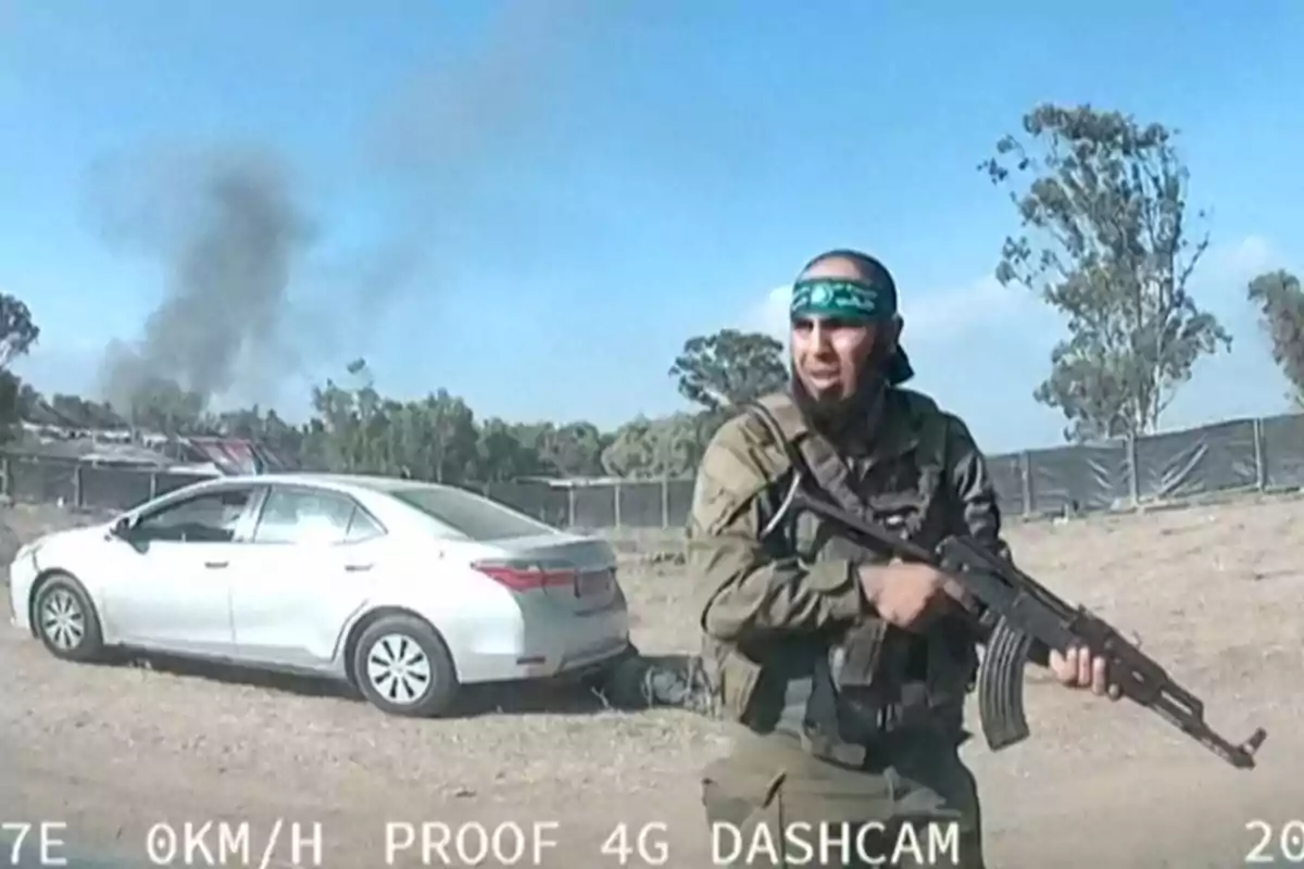 Hombre armado con uniforme militar y banda verde en la cabeza caminando junto a un auto blanco estacionado en un camino de tierra con humo negro al fondo y árboles alrededor