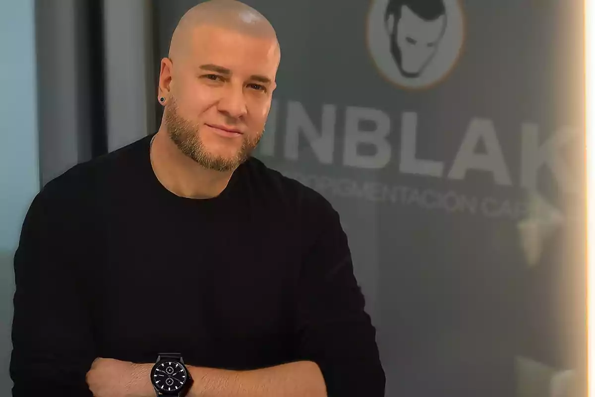 Hombre calvo con barba y camiseta negra posando frente a un fondo con un logo y texto desenfocado