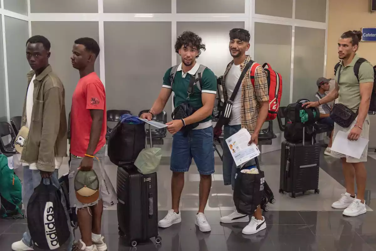 Cinco hombres jóvenes con equipaje hacen fila en una sala de espera, algunos sostienen documentos y mochilas, todos visten ropa casual y parecen estar en un entorno de oficina o terminal.