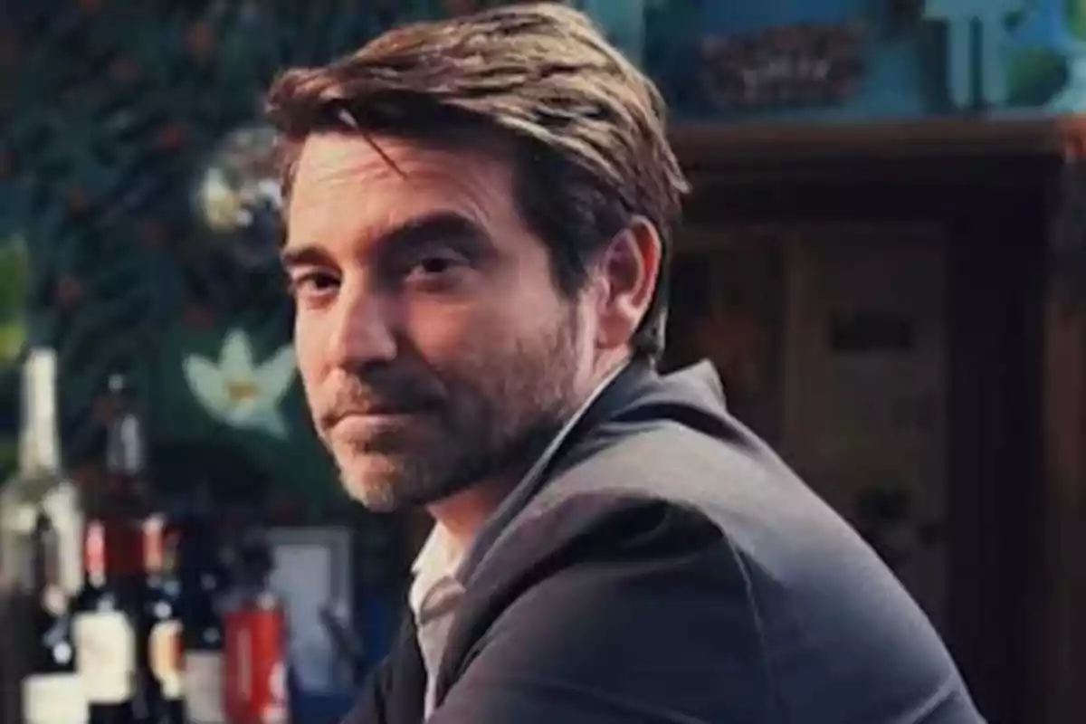 Hombre de cabello castaño y barba corta con saco oscuro sentado en un bar mirando hacia la cámara