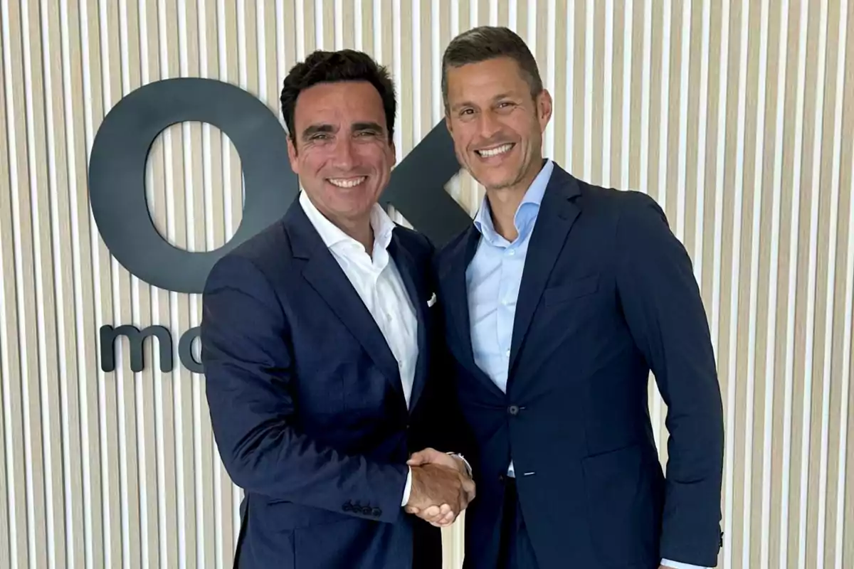 Dos hombres de negocios sonríen y se dan la mano frente a una pared con un logotipo de empresa.