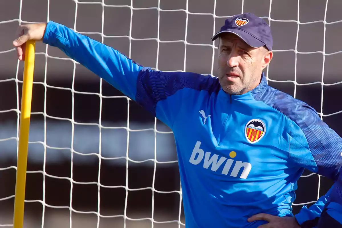 Entrenador del Valencia CF con ropa deportiva azul apoyado en un poste amarillo frente a una portería de fútbol