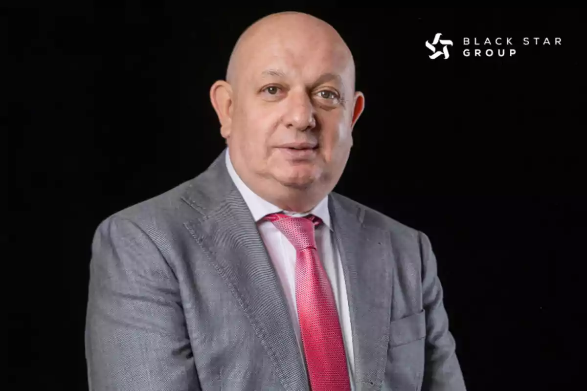 Hombre calvo con traje gris y corbata rosa posando frente a un fondo negro con el logo de Black Star Group en la esquina superior derecha