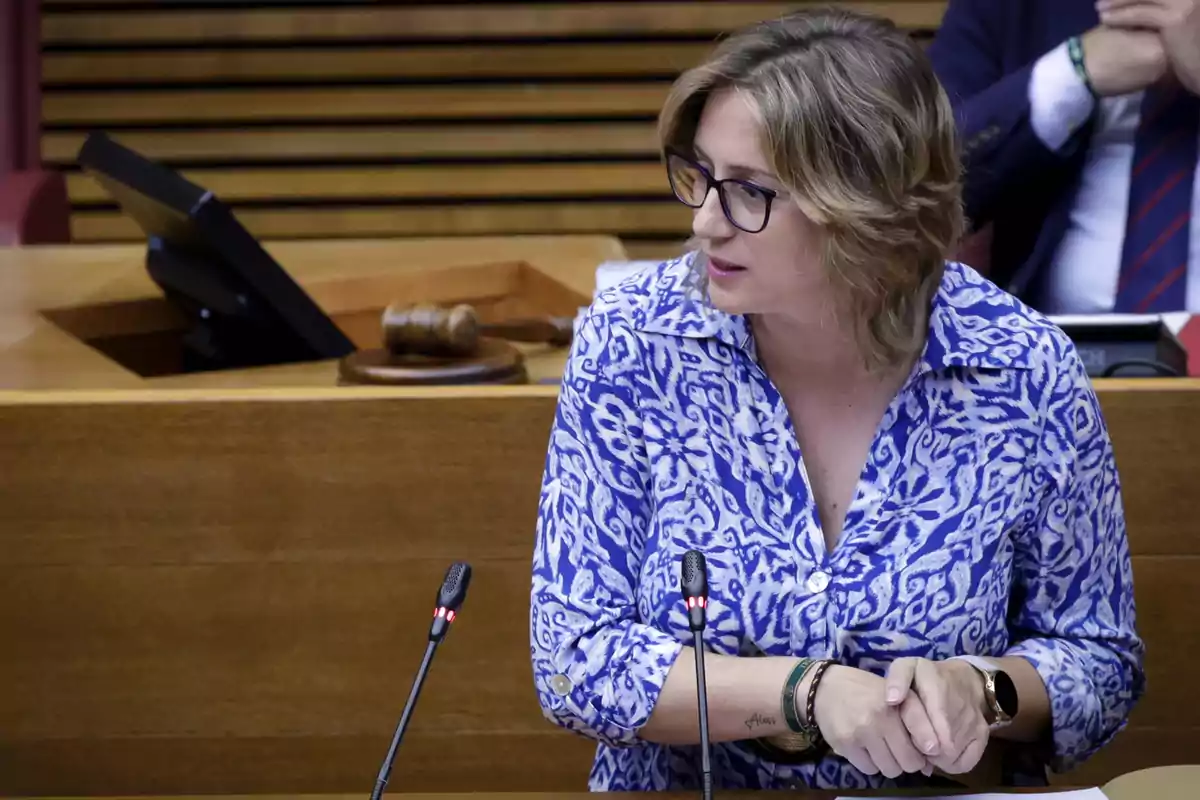 Mujer con gafas y camisa azul hablando frente a dos micrófonos en una sala de reuniones o parlamento Mujer con gafas y camisa azul hablando frente a dos micrófonos en una sala de reuniones o parlamento