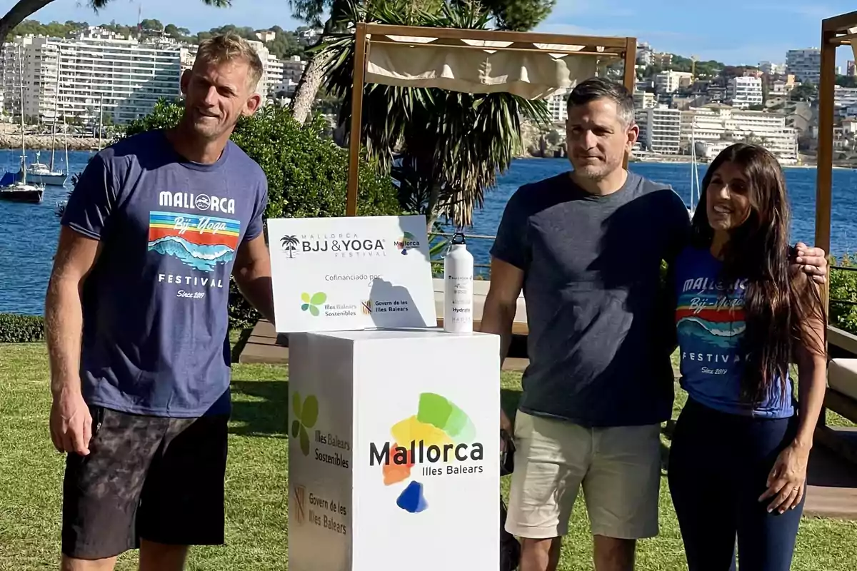 Tres personas posan al aire libre junto a un cartel del Mallorca BJJ & Yoga Festival con el mar y edificios al fondo en un día soleado