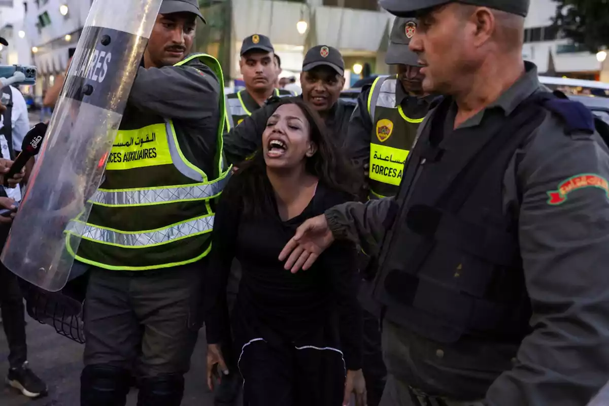 Una mujer grita mientras es detenida por varios policías con chalecos reflectantes y escudos en una calle concurrida. Una mujer grita mientras es detenida por varios policías con chalecos reflectantes y escudos en una calle concurrida.