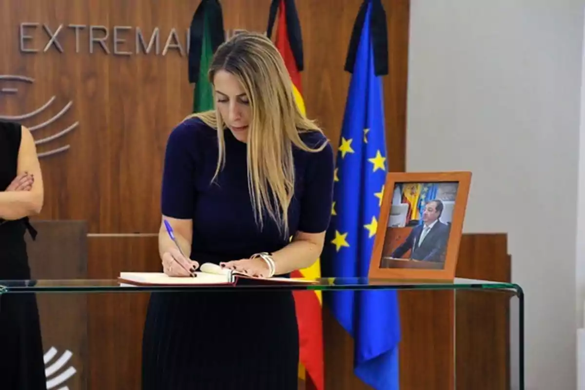 Mujer rubia firmando un libro sobre una mesa de cristal junto a un retrato enmarcado de un hombre con traje y corbata, al fondo hay banderas y una pared de madera Mujer rubia firmando un libro sobre una mesa de cristal junto a un retrato enmarcado de un hombre con traje y corbata, al fondo hay banderas y una pared de madera