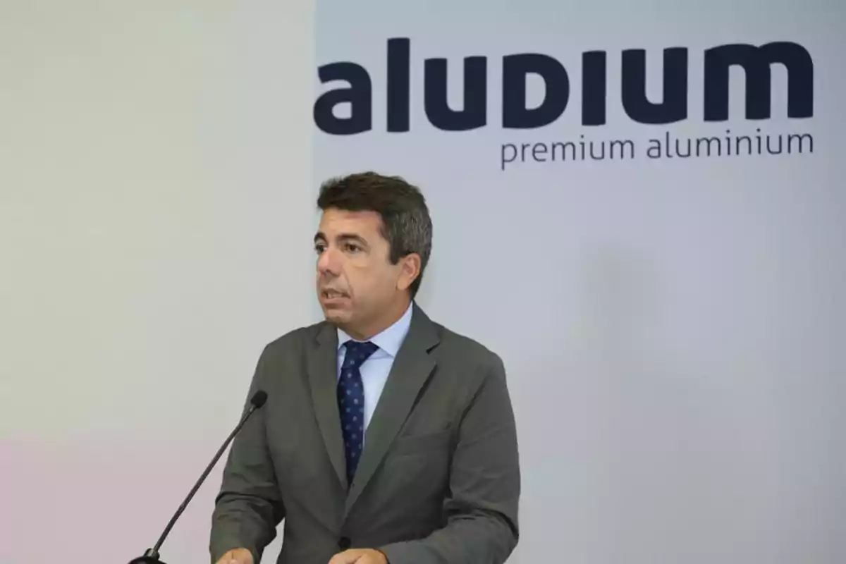 Hombre de traje hablando frente a un micrófono con un cartel de la empresa Aludium al fondo