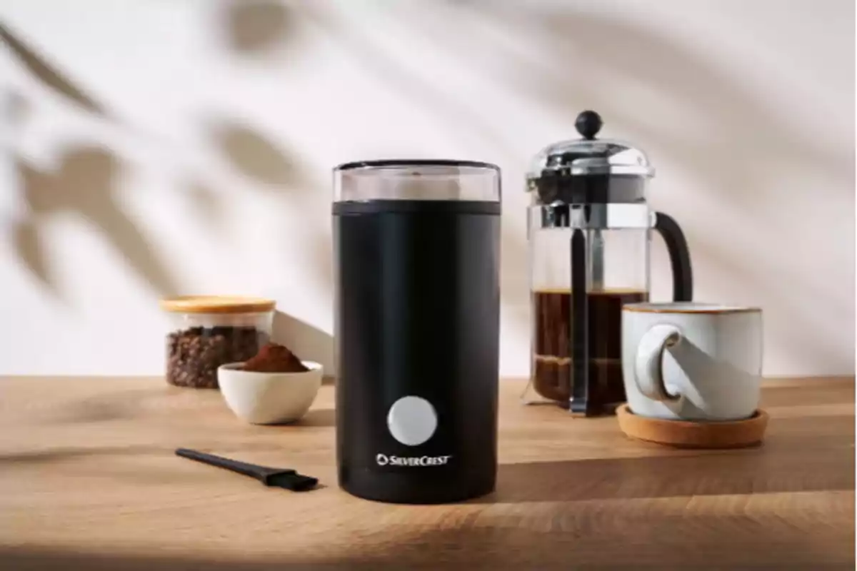 Molino de café eléctrico negro sobre una mesa de madera junto a una prensa francesa, una taza de café, un frasco con granos y un recipiente con café molido