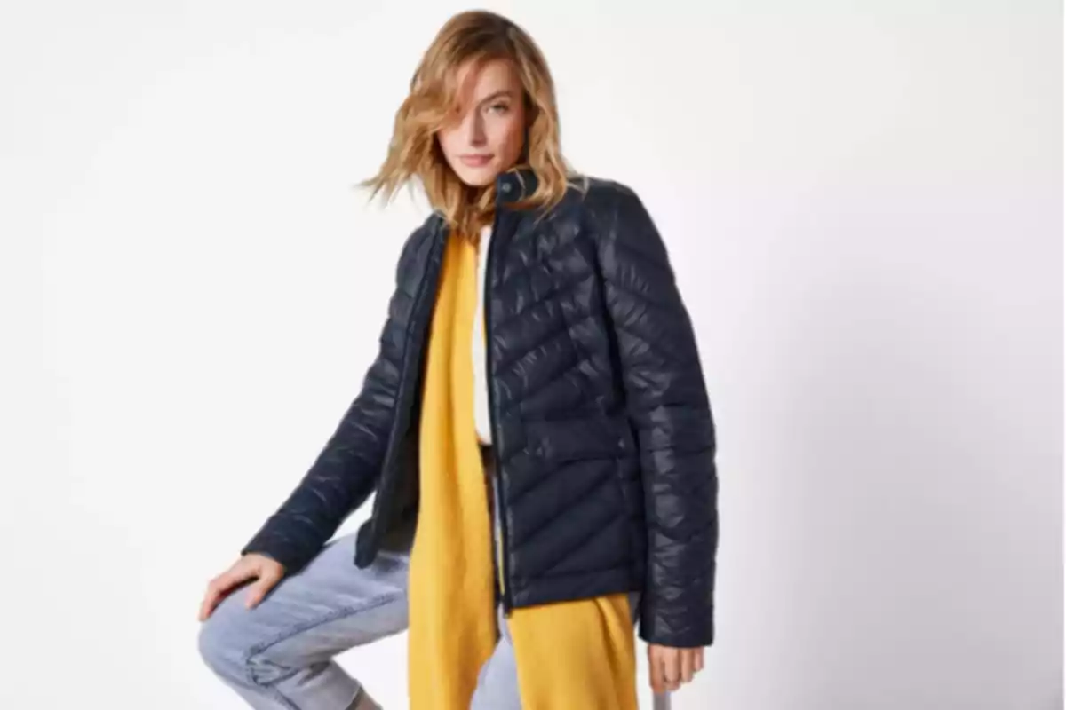 Mujer con chaqueta acolchada azul marino, bufanda amarilla y pantalón claro posando frente a un fondo blanco