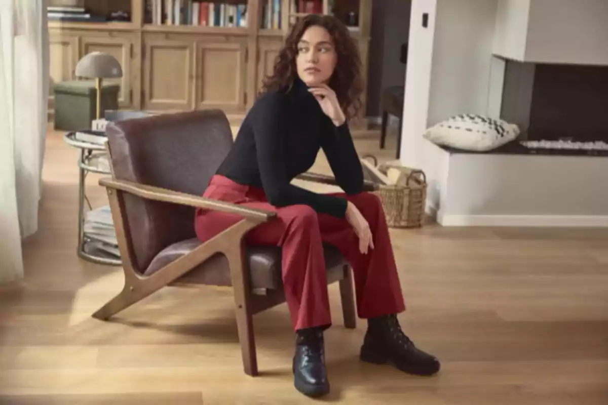 Mujer sentada en una silla de madera con asiento de cuero, lleva un suéter negro de manga larga, pantalones rojos y botas negras, en una sala con piso de madera y decoración moderna