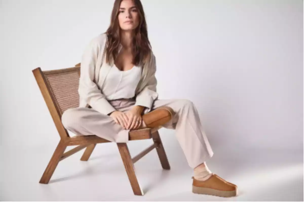 Mujer sentada en una silla de madera con ropa beige y zapatos marrones en un fondo claro