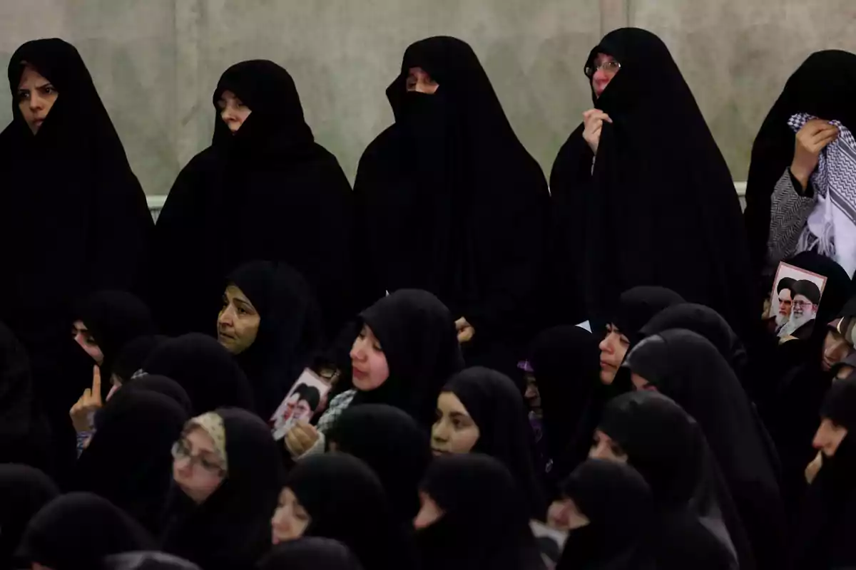 Grupo de mujeres vestidas con chador negro reunidas en un espacio interior algunas sostienen fotografías y muestran expresiones serias o de tristeza Grupo de mujeres vestidas con chador negro reunidas en un espacio interior algunas sostienen fotografías y muestran expresiones serias o de tristeza