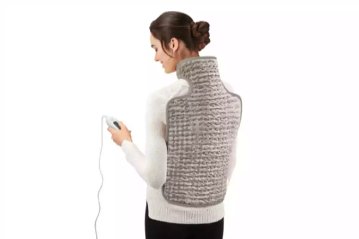 Mujer de espaldas usando una manta térmica eléctrica en la espalda y cuello mientras sostiene el control en la mano Mujer de espaldas usando una manta térmica eléctrica en la espalda y cuello mientras sostiene el control en la mano
