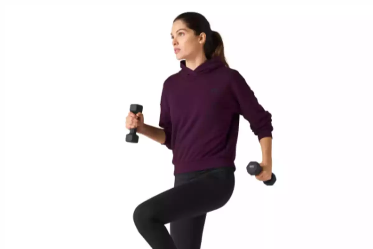 Mujer joven haciendo ejercicio con pesas pequeñas vestida con sudadera morada y pantalones negros sobre fondo blanco