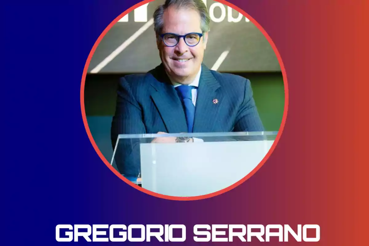 Hombre de cabello canoso y gafas azules sonríe mientras está de pie detrás de un atril con fondo degradado en tonos azul y rojo y el nombre Gregorio Serrano en la parte inferior.