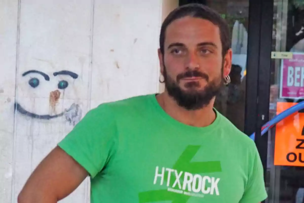 Hombre con barba y camiseta verde de HTX Rock de pie junto a una pared con un dibujo de una cara sonriente