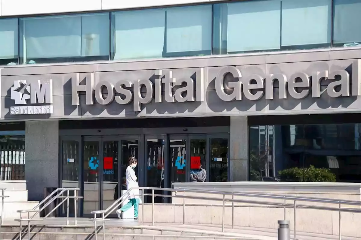 Fachada de un hospital general con una persona con bata médica y mascarilla caminando frente a la entrada principal Fachada de un hospital general con una persona con bata médica y mascarilla caminando frente a la entrada principal