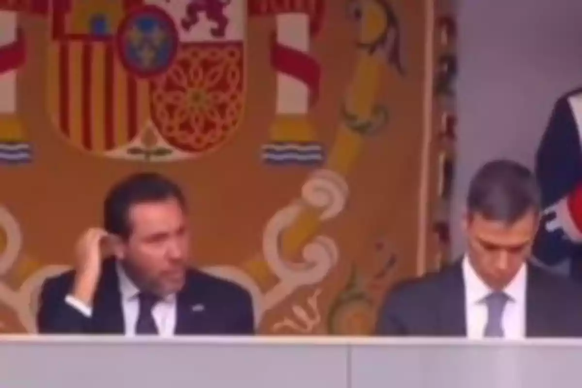 Dos hombres con traje están sentados frente a un escudo grande, uno se rasca la cabeza y el otro mira hacia abajo