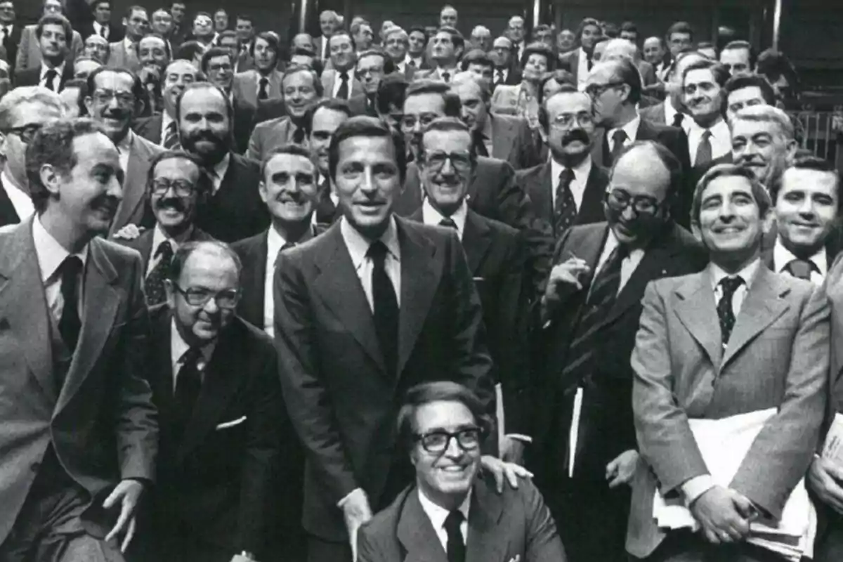 Grupo de hombres vestidos con trajes formales sonríen y posan juntos en una fotografía en blanco y negro