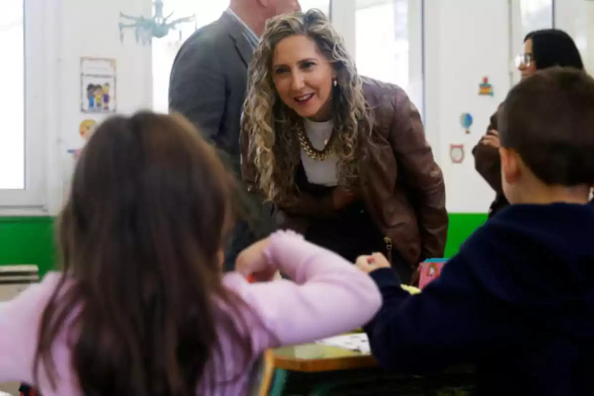 Mujer de cabello rizado y chaqueta marrón sonriendo e inclinándose hacia dos niños que están sentados en un aula escolar Mujer de cabello rizado y chaqueta marrón sonriendo e inclinándose hacia dos niños que están sentados en un aula escolar
