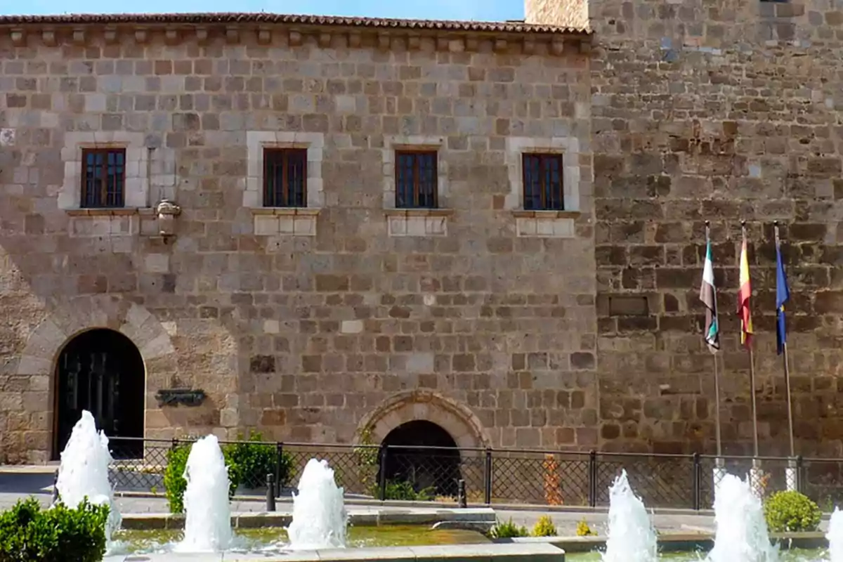 Fachada de un edificio histórico de piedra con varias ventanas, una puerta de arco, una fuente con chorros de agua en primer plano y tres banderas ondeando a la derecha. Fachada de un edificio histórico de piedra con varias ventanas, una puerta de arco, una fuente con chorros de agua en primer plano y tres banderas ondeando a la derecha.