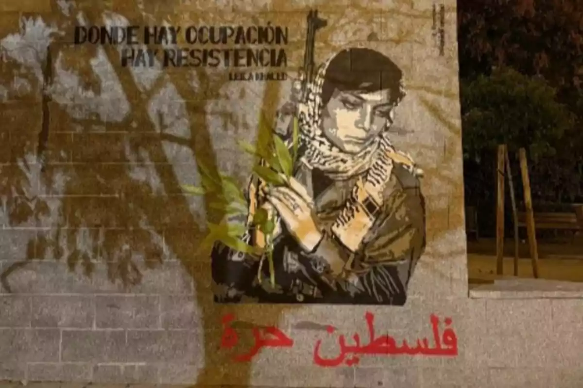Mural en una pared que muestra a una persona con un pañuelo palestino, sosteniendo una rama de olivo y un arma, acompañado de frases en español y árabe relacionadas con la resistencia y Palestina