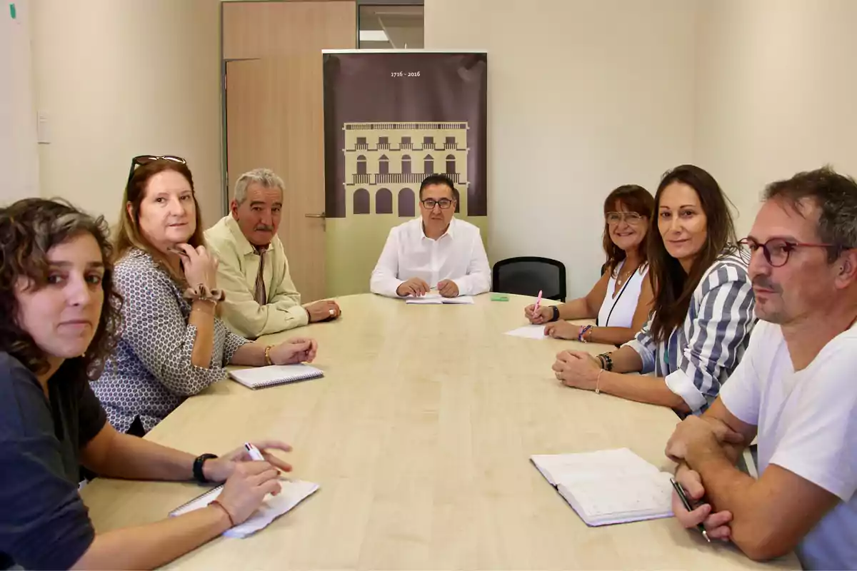 Siete personas sentadas alrededor de una mesa de reuniones en una oficina, algunas tomando notas y mirando hacia la cámara