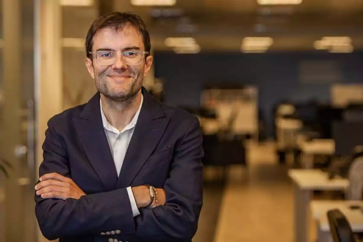 Hombre con gafas y saco azul marino sonriendo y posando con los brazos cruzados en una oficina moderna iluminada