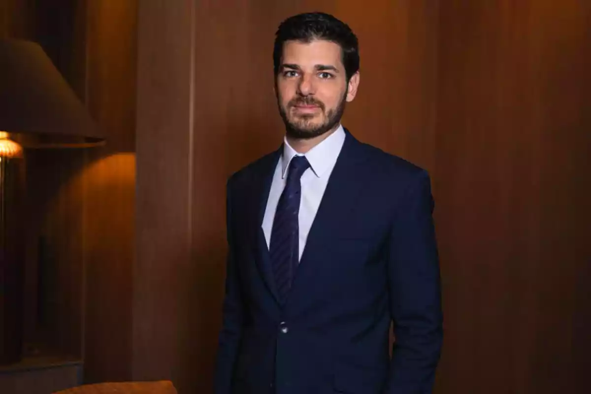 Hombre joven con barba y cabello oscuro vistiendo un traje azul marino y corbata, posando en un ambiente interior elegante con paredes de madera.